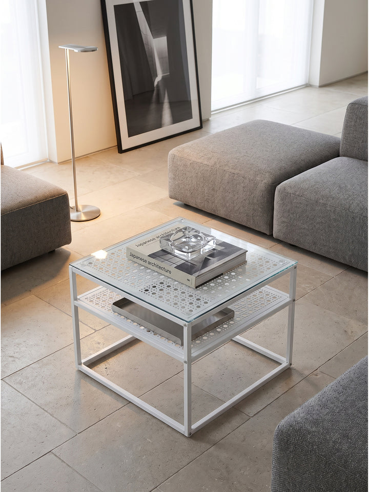 Quadratischer Glastisch mit Ablagefläche von oben betrachtet neben einem hellgrauen Loungesofa und einer Aluminium-Stehleuchte._
