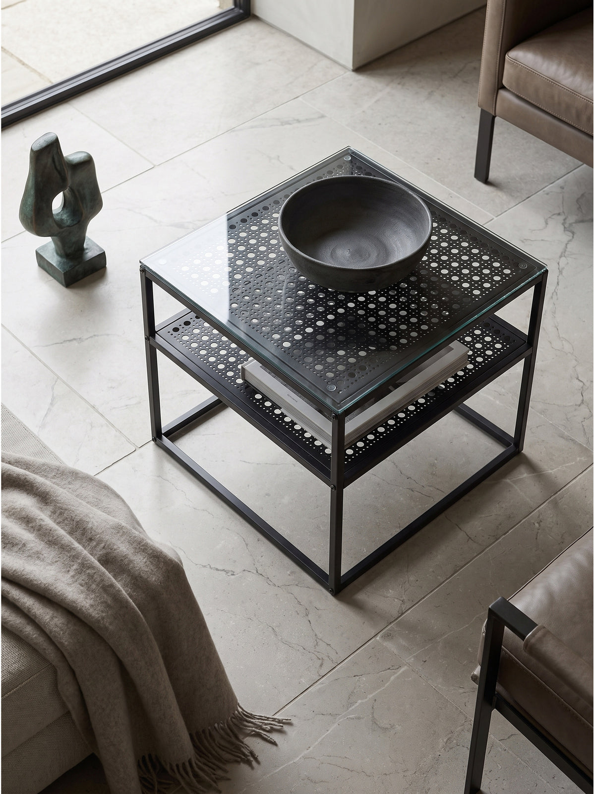 Aufsicht auf modernen Glastisch mit schwarzem Metallgestell und gelochter Ablage neben minimalistischem Sofa_