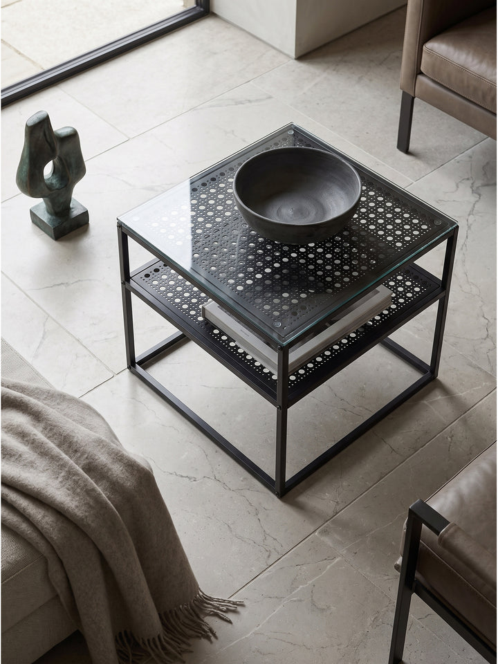 Aufsicht auf modernen Glastisch mit schwarzem Metallgestell und gelochter Ablage neben minimalistischem Sofa_