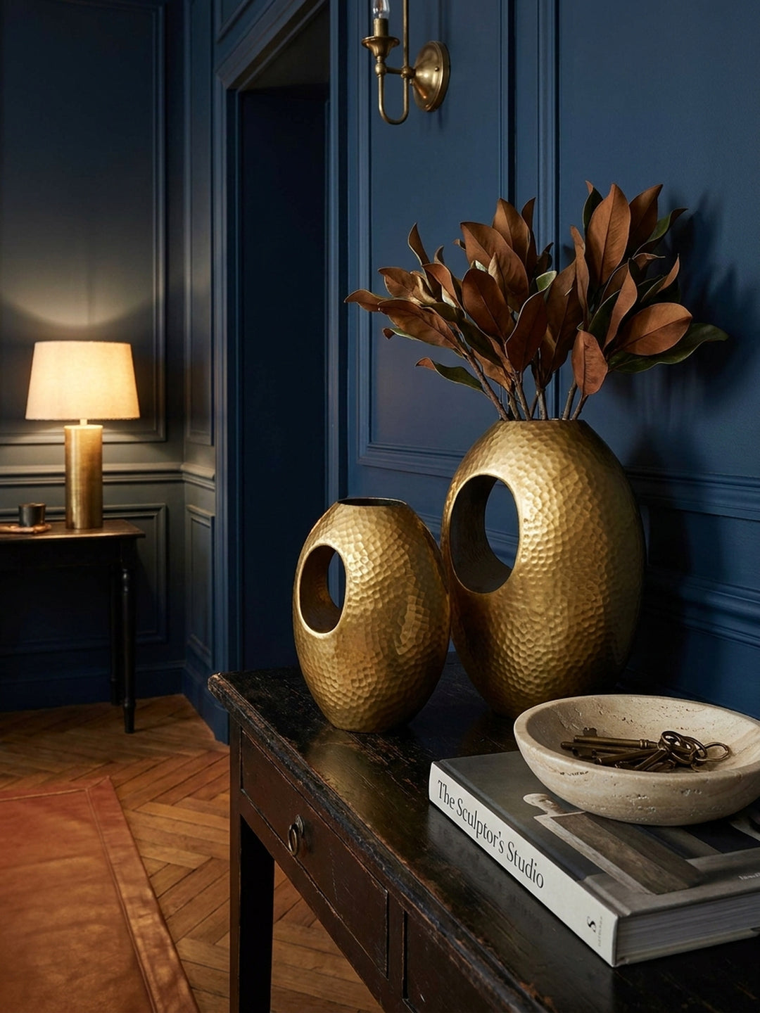 Stilvolles Deko-Vignette mit der modernen Blumenvase in Gold, gefüllt mit Eukalyptuszweigen, neben einer brennenden Kerze und einem Stapel Bücher auf einem Sideboard._