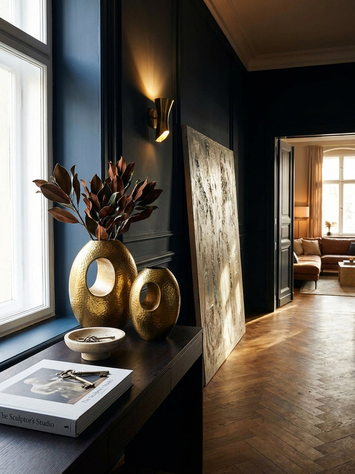 Das zweiteilige Vasen-Set in Gold harmoniert auf einem dunklen Eichenholz-Sideboard neben einer Tischlampe aus Messing und einer schwarzen Marmorschale._
