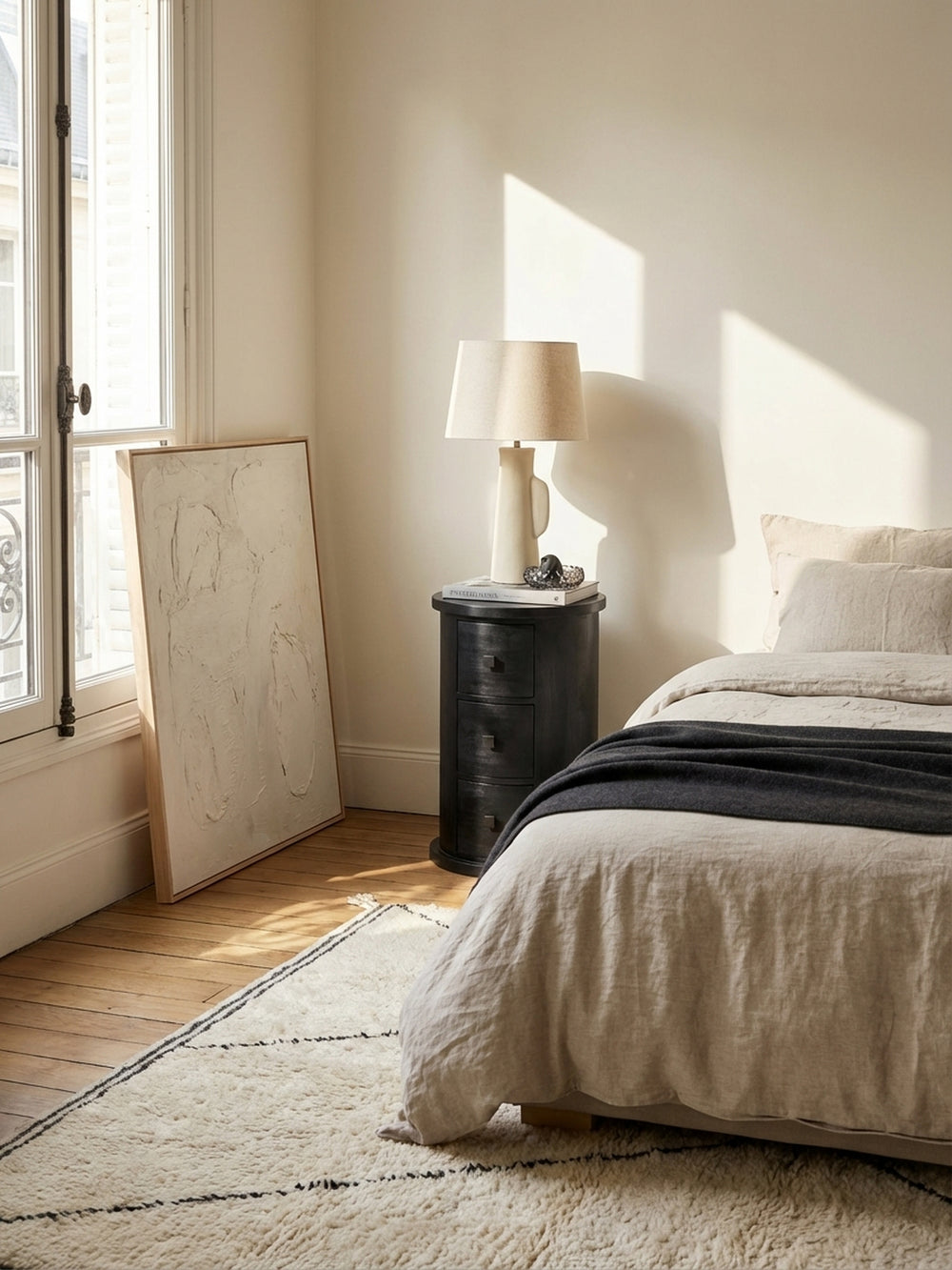 Der runde Nachttisch aus massivem Mangoholz in Schwarz steht in einem hellen Schlafzimmer auf einem flauschigen Beni Mrirt Teppich neben einem Leinenbett._