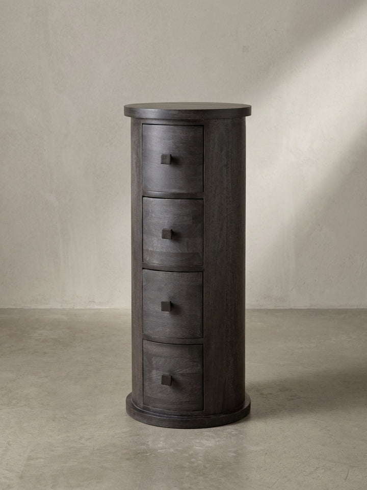 Dreiviertelansicht des zylinderförmigen Beistellschranks aus massivem Mangoholz, der die Tiefe der geschwungenen Schubladen und die schlanke Silhouette hervorhebt._