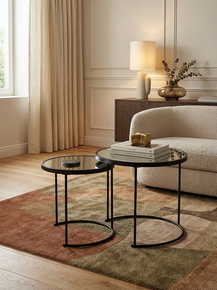 Modernes Satztisch-Set aus schwarzem Metall und Spiegelglas, elegant inszeniert neben einem grauen Sofa auf einem texturierten Teppich im Wohnzimmer._
