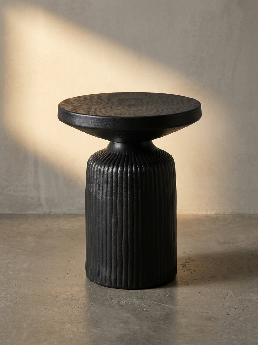 Schwarzer runder Anstelltisch aus Metall in der 3/4-Ansicht, der die Lamellen-Optik des Fußes und die massive Tischplatte hervorhebt._