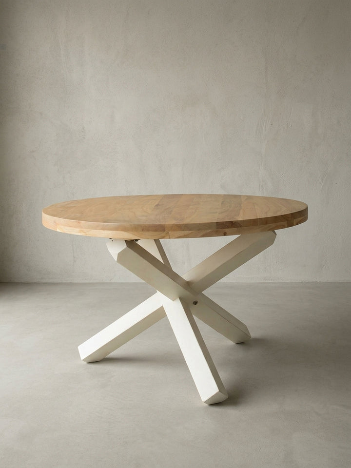 Freigestellter runder Esstisch im modernen Landhausstil. Die frontale Ansicht zeigt die braune Massivholzplatte aus Akazie und das markante dreibeinige Kreuzgestell in Cremeweiß._