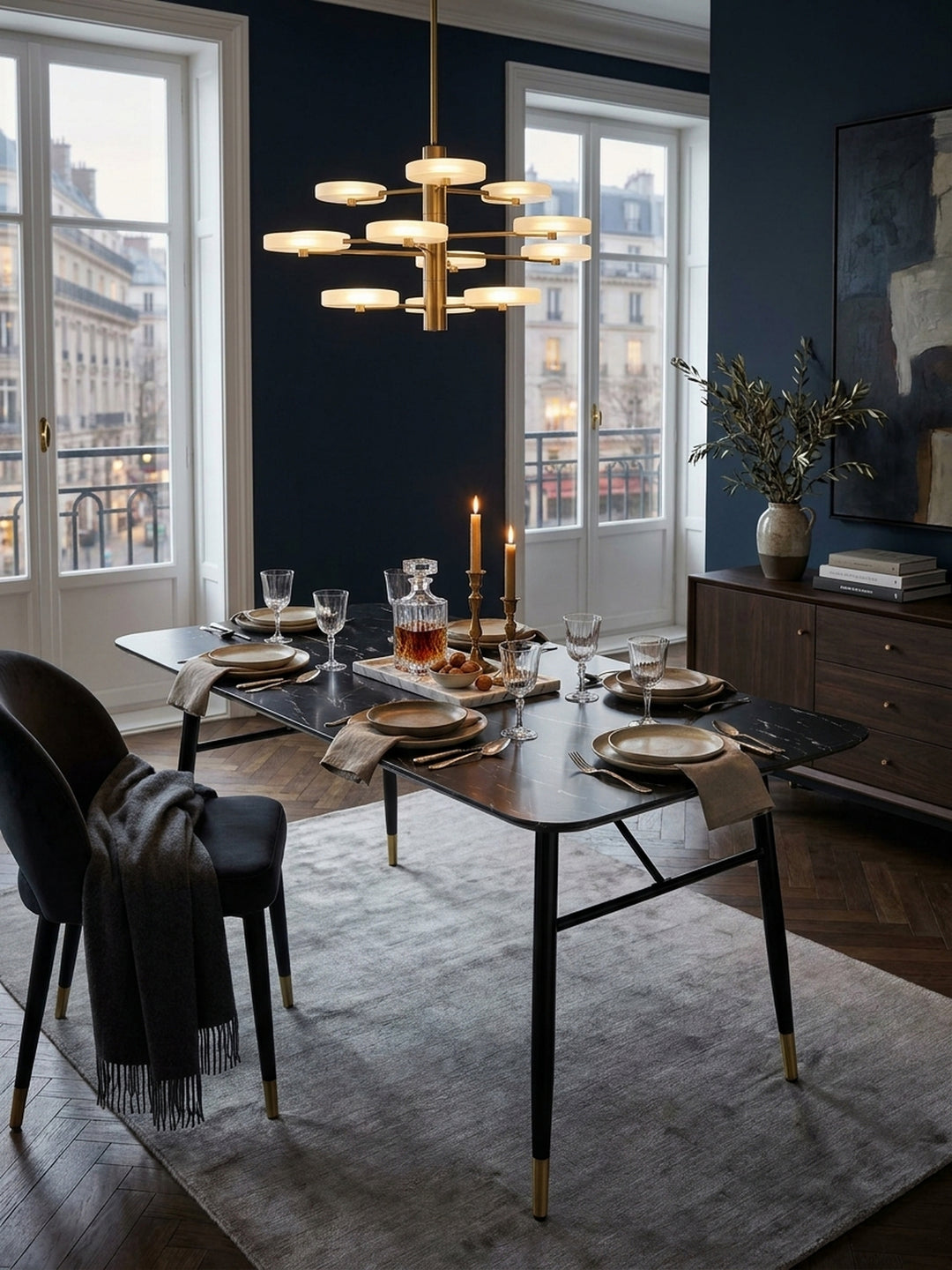 Moderner Esstisch 140x80 cm in schwarzer Marmoroptik mit goldenen Fußkappen arrangiert in einem eleganten Esszimmer mit tiefblauer Wand und dunklem Fischgrätparkett._