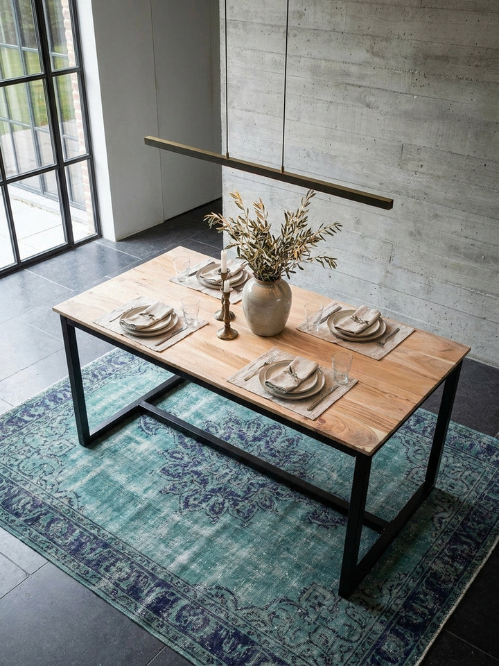 Draufsicht auf den rechteckigen Esszimmertisch aus Akazie im Industrial Style, umgeben von eleganten Polsterstühlen auf einem blauen Vintage Teppich im weitläufigen Wohnbereich._