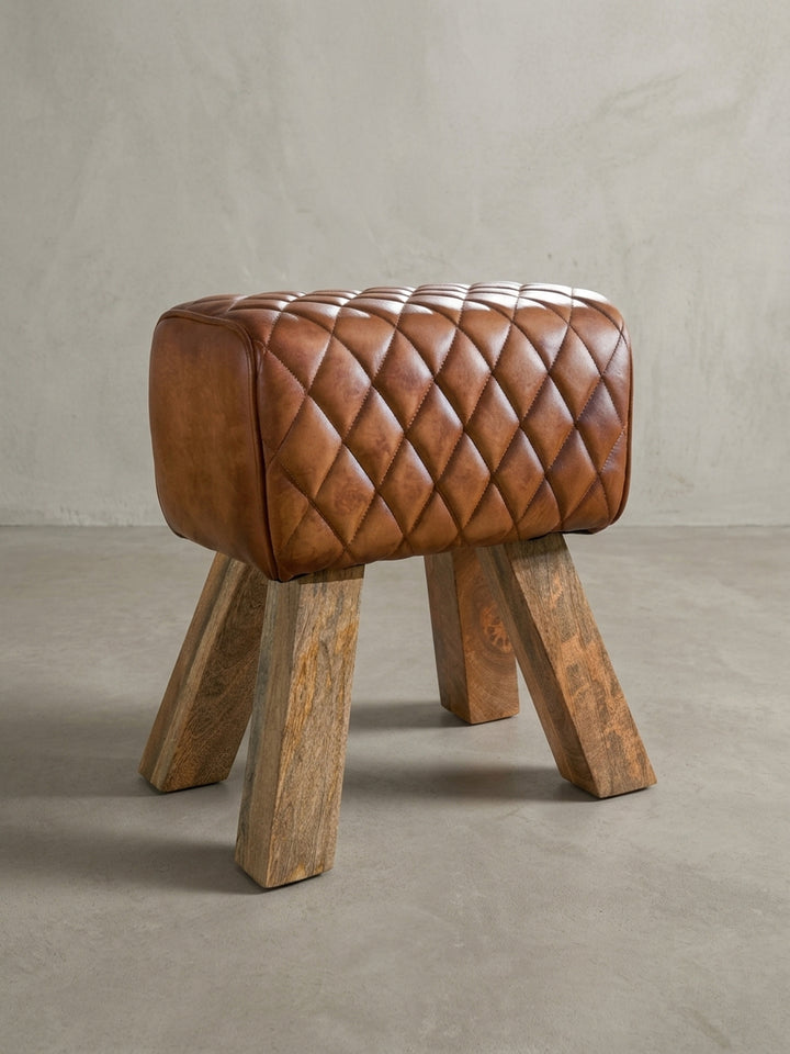 Design-Hocker in Turnbock-Form, Dreiviertelansicht, zeigt das Zusammenspiel von gestepptem Leder und den schrägen Holzfüßen des Fußhockers._