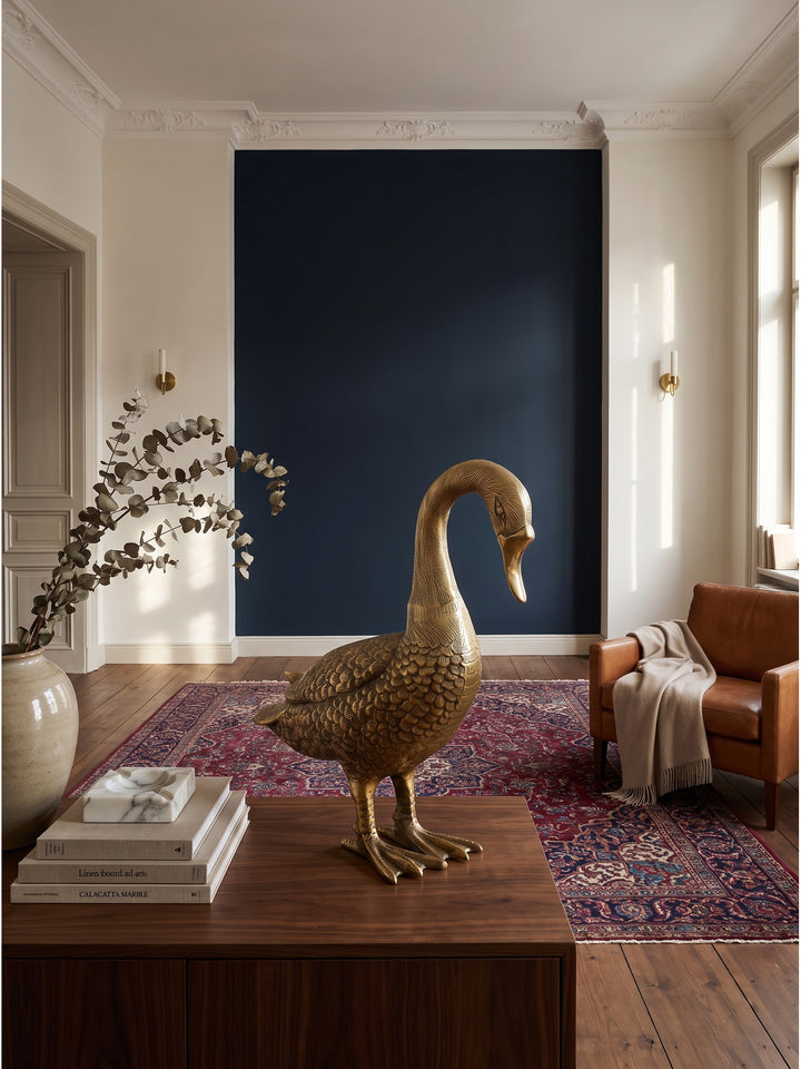 Große Dekofigur Gans aus golden lackiertem Aluminium 46x19x42 cm als elegantes Statement-Piece auf einem dunklen Walnuss-Sideboard in einem klassisch modernen Wohnzimmer vor nachtblauer Wand._