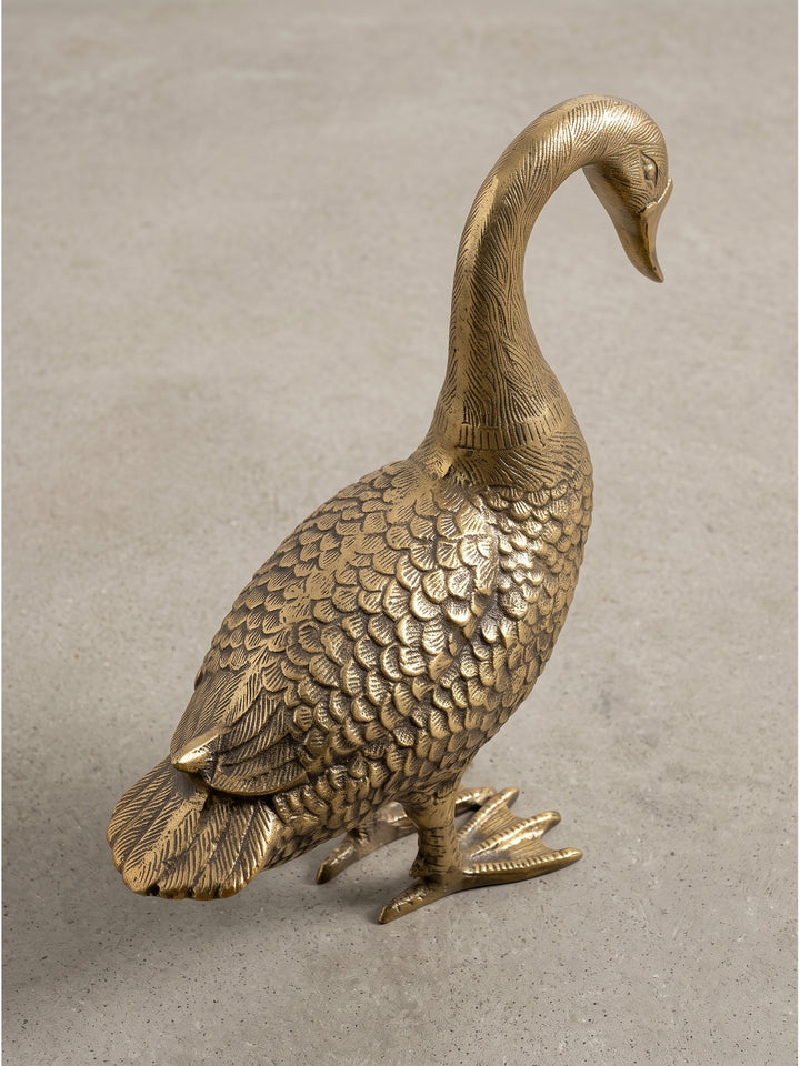 Moderne Tischdekoration in Form einer 46 cm langen Gans aus lackiertem Aluminium im schrägen Winkel, die warmen patinierten Goldtöne verleihen dem Wohnaccessoire einen fühlbar luxuriösen Charakter._
