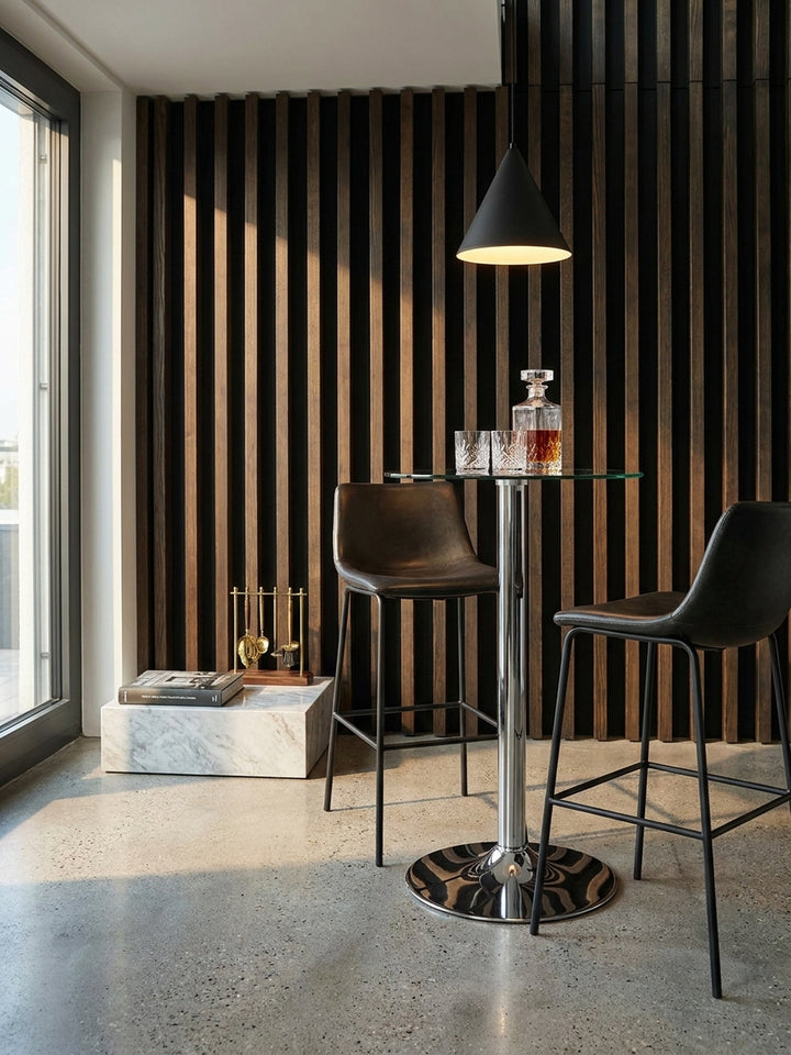 Der elegante Chrom-Bistrotisch arrangiert mit zwei dunkelbraunen minimalistischen Barhockern vor einer bodentiefen Stahlrahmen-Trennwand im privaten Barbereich._