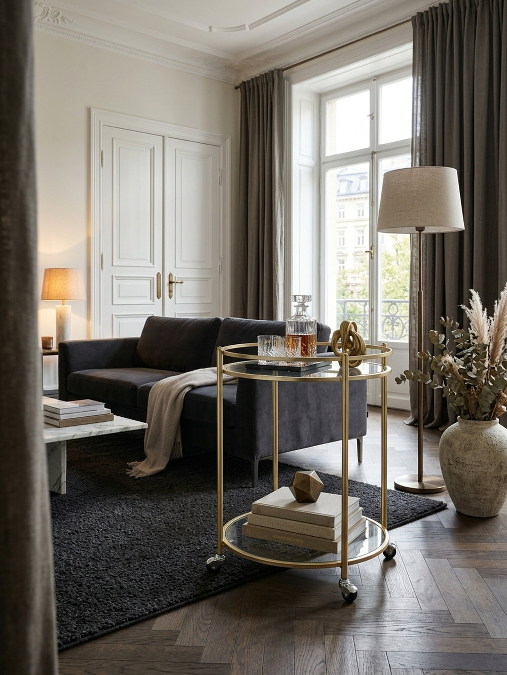 Eleganter runder Servierwagen in mattem Gold mit zwei klaren Glasplatten, stilvoll als Barwagen im luxuriösen Wohnzimmer mit Fischgrätparkett und dunklem Bouclé-Teppich inszeniert._