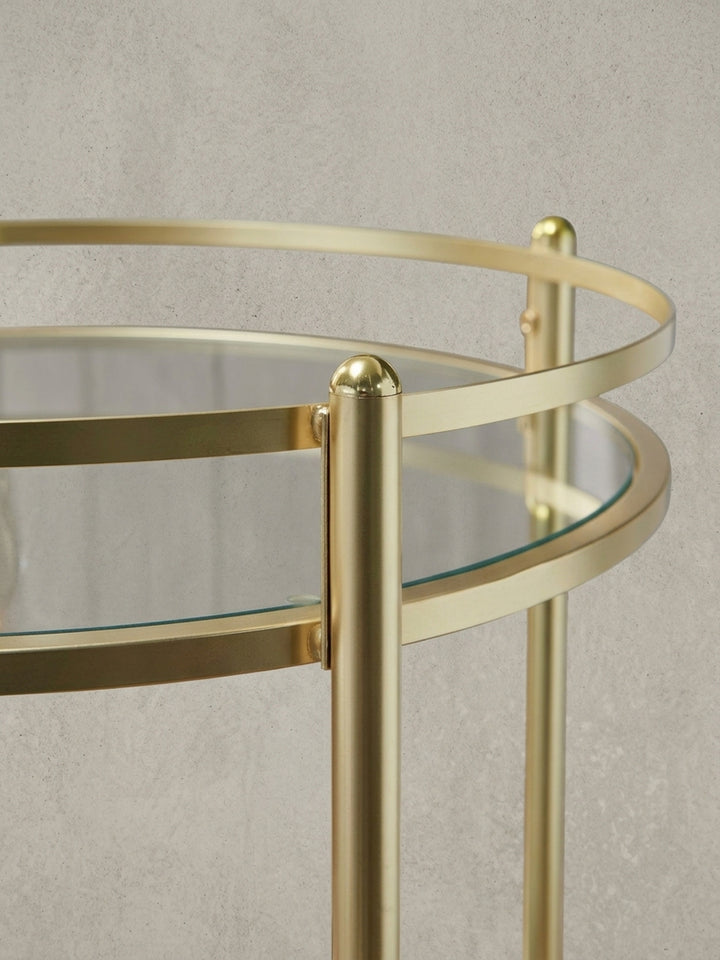 Alternative Perspektive des goldenen Rollwagens mit klaren Glasplatten. Der 57 cm breite Serviertisch beweist seine zeitlose Eleganz als vielseitiges Möbelstück für das Wohnzimmer._