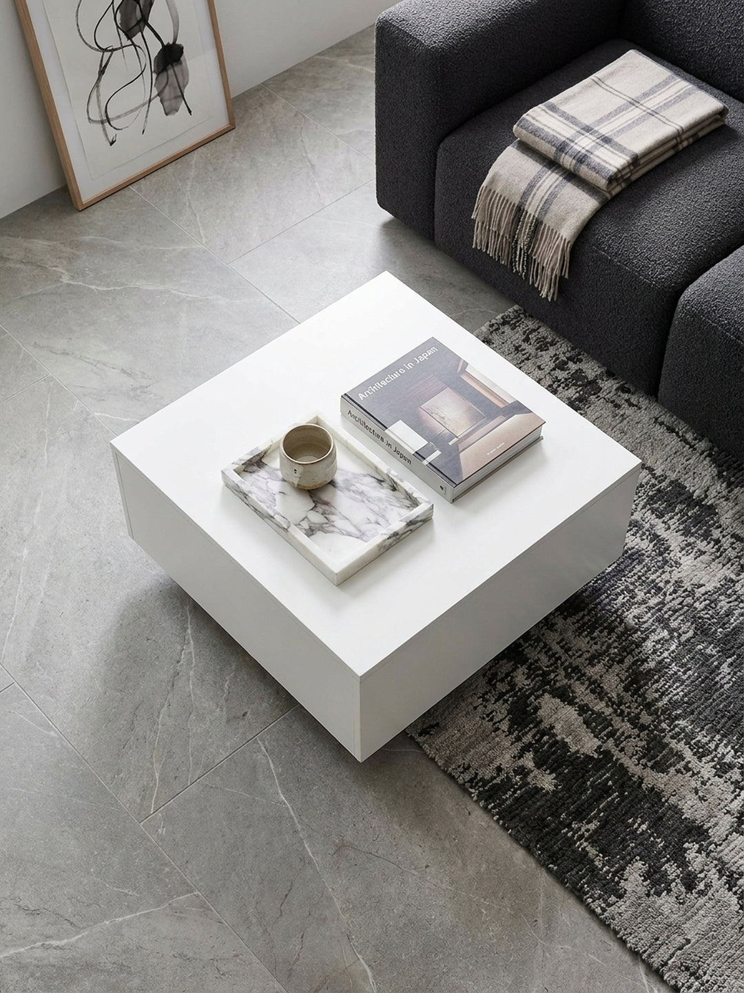 Quadratischer Loungetisch mit silbernem Sockel von oben betrachtet, mittig platziert zwischen einem modularen grauen Bouclé-Sofa und Architektur-Bildband._
