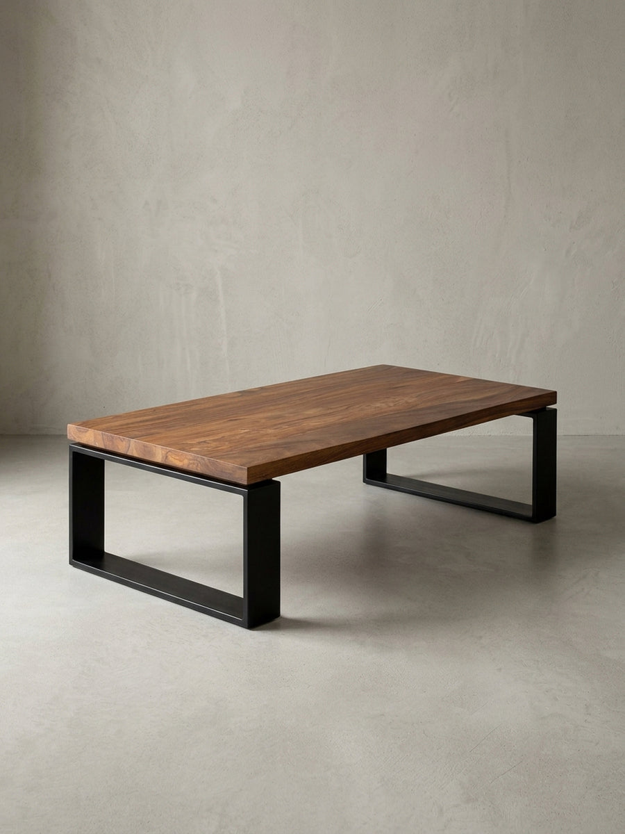 Massiver Couchtisch 115x35x60 cm im Studio. Die braune Sheeshamholz-Platte ruht auf zwei robusten, matt schwarzen Eisenkufen und zeigt ein klares, industrielles Design._