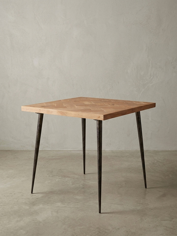 Frontalansicht des quadratischen Esstisches 80x80x76 cm. Die massive Akazienholzplatte mit Fischgrätmuster harmoniert perfekt mit den dunklen, konischen Metallbeinen im Industrial-Design._