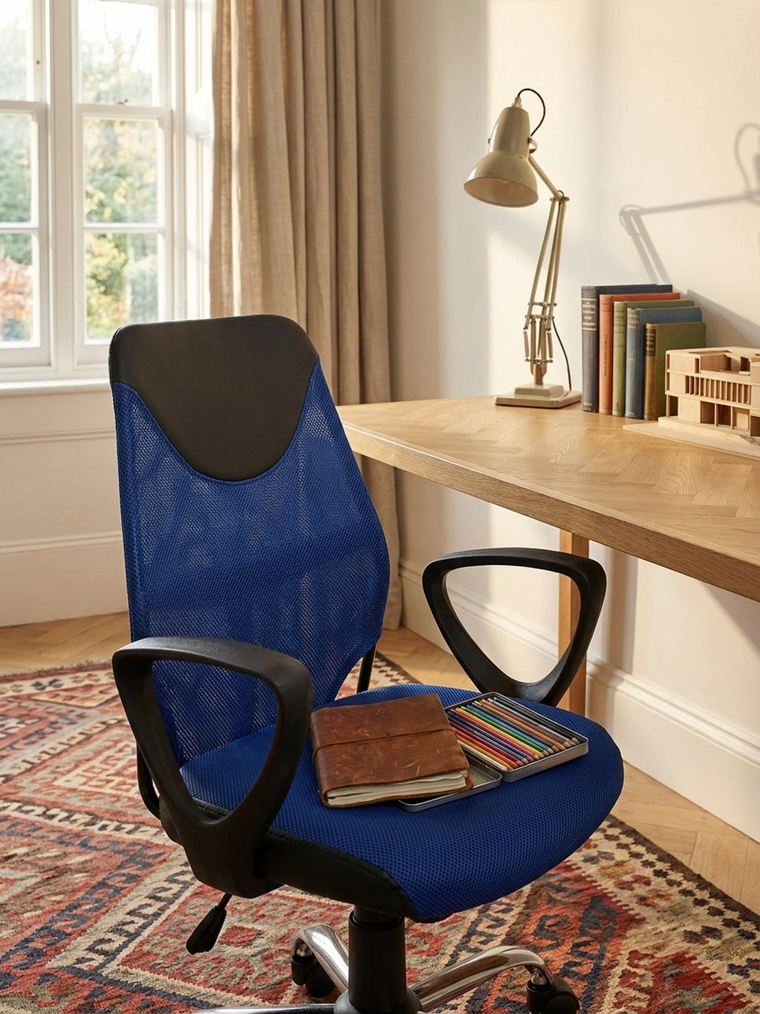 Blaue Sitzfläche des höhenverstellbaren Jugendstuhls an einem aufgeräumten Schreibtisch mit Skizzenbüchern und einer matten weißen Schreibtischlampe._