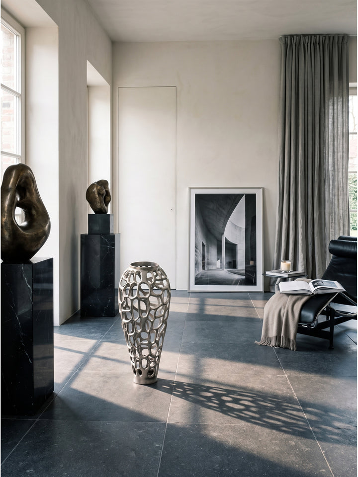 Silberne Bodenvase aus Aluminium mit organischer Lochstruktur 60cm hoch in einem minimalistischen Wohnzimmer mit großen Fenstern und dunklem Natursteinboden_