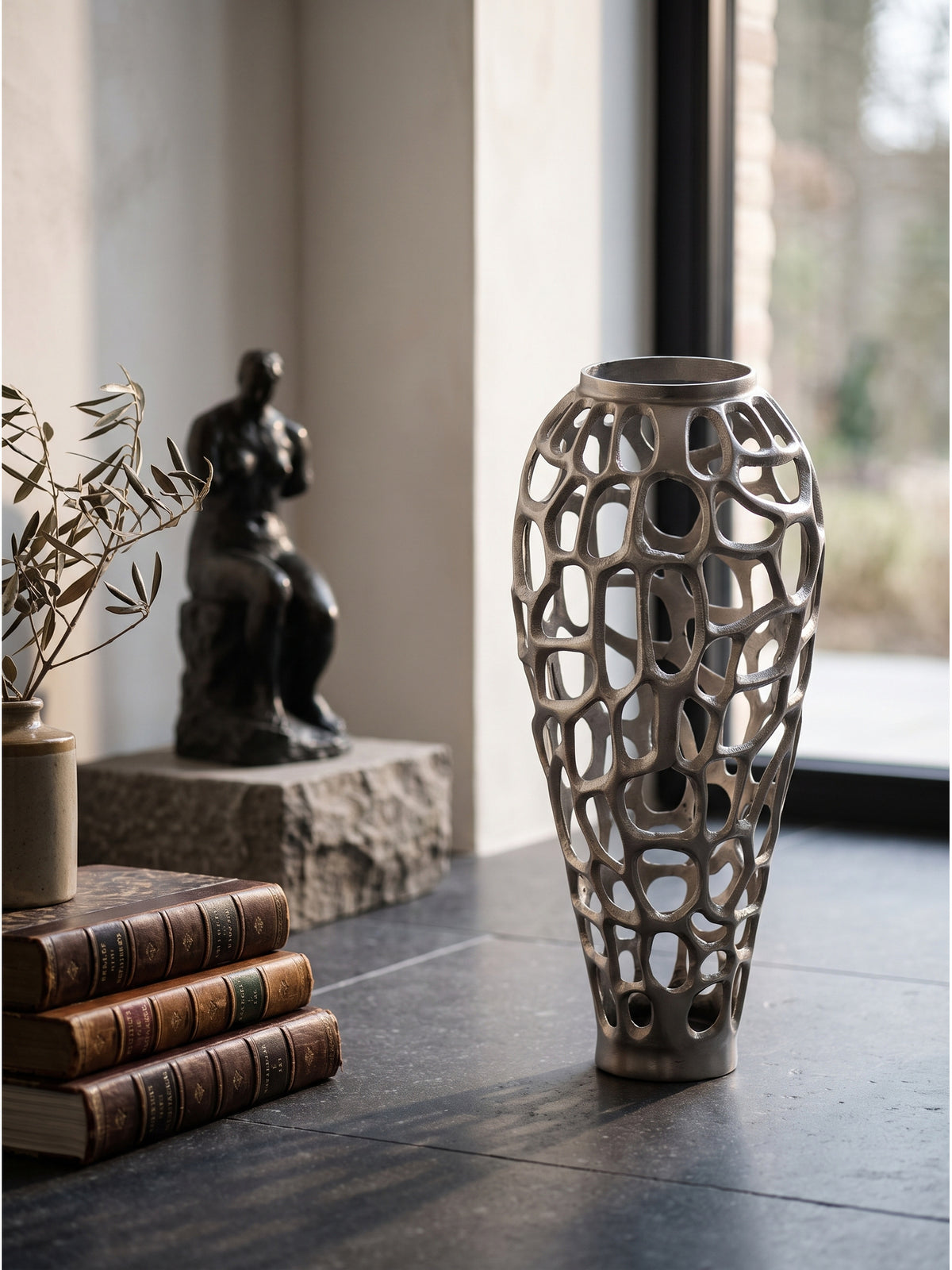 Große Bodenvase aus Aluminium in Silber als skulpturales Objekt dekoriert mit Trockenblumen neben einem flachen schwarzen Marmorpodest im Eingangsbereich_