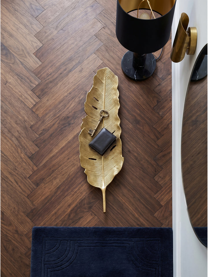 Aufsicht auf die längliche goldene Blattschale aus Aluminium, stilvoll drapiert mit Ringen und einer Sonnenbrille, auf einer edlen Holzkonsole im eleganten Art-Deco Wohnzimmer._