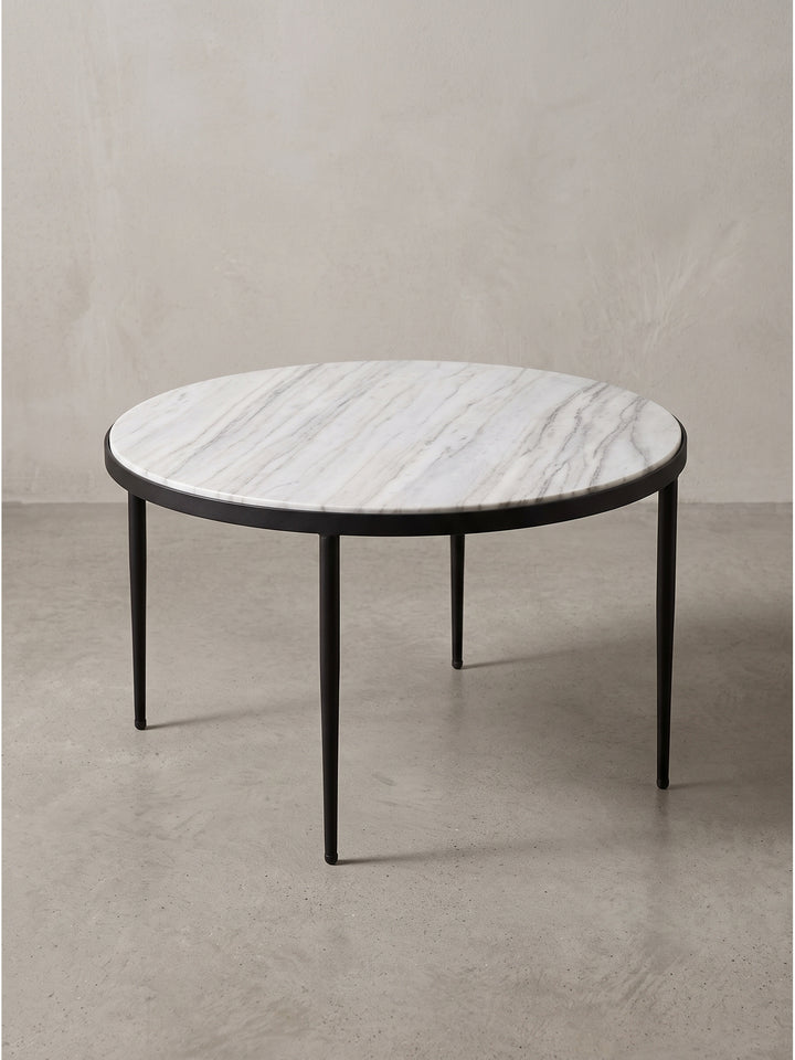 Freigestellter Couchtisch mit weiß-grauer Marmorplatte und drei spitz zulaufenden schwarzen Metallbeinen vor neutralem Hintergrund._