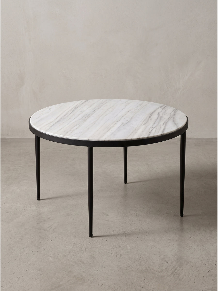 Freigestellter Couchtisch mit weiß-grauer Marmorplatte und drei spitz zulaufenden schwarzen Metallbeinen vor neutralem Hintergrund._