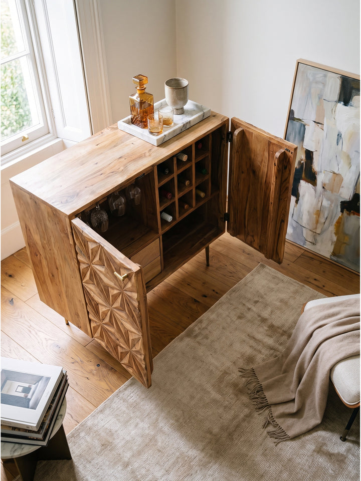 Hausbar aus massivem Akazienholz mit 3D Muster und goldfarbenen Beinen vor strukturierter cremefarbener Wand arrangiert_