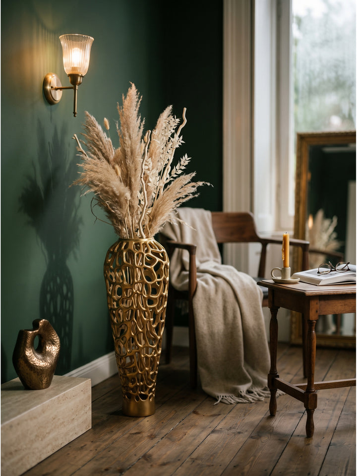 Trockenblumen und helle Pampaswedel arrangiert in einer bauchigen goldenen Dekovase auf dem Fußboden neben einem Nussbaum-Sideboard_