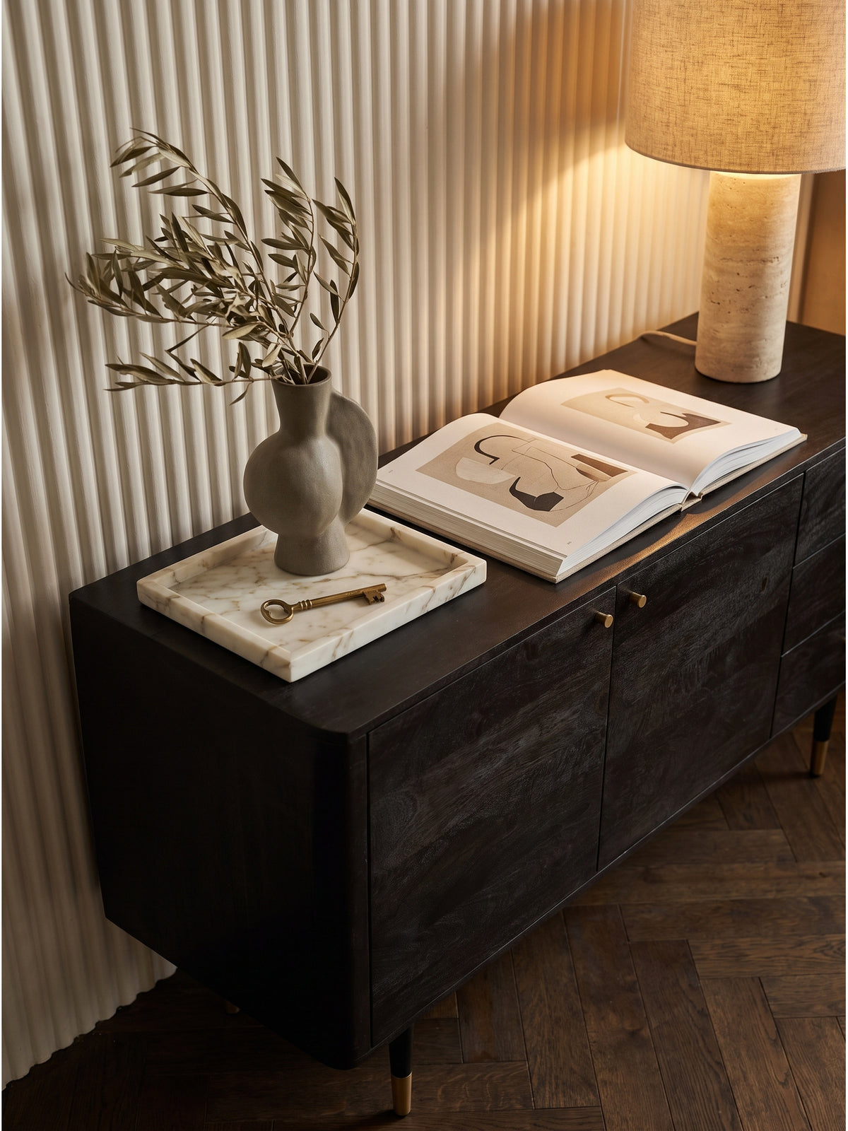 Dekoriertes Wohnzimmer-Sideboard aus Massivholz mit einer schweren Travertin-Tischlampe und getrockneten Olivenzweigen in rauer Keramik.