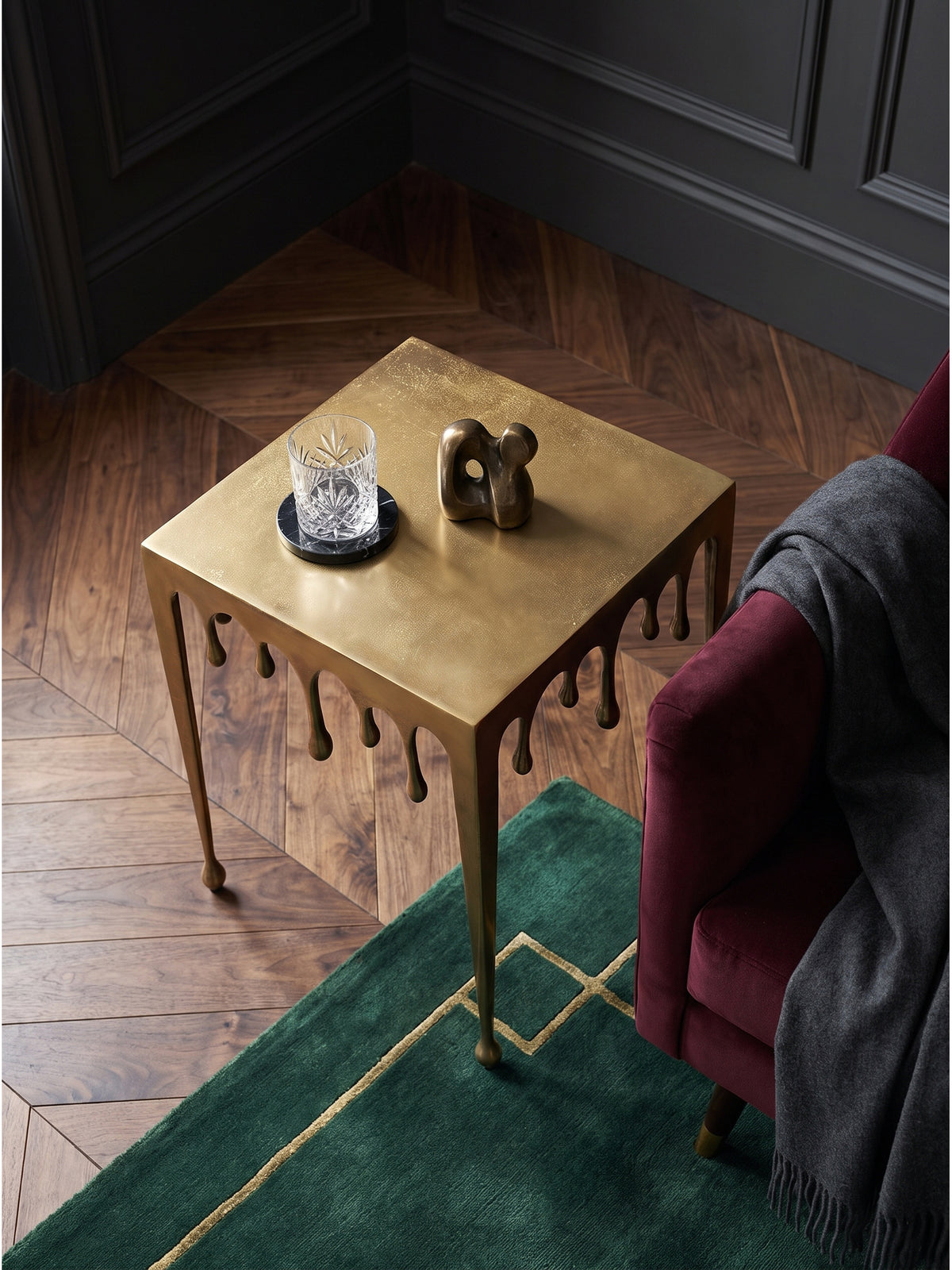 Draufsicht auf den quadratischen goldenen Metalltisch mit Tropfen-Design neben einem samtigen Sessel auf dunklem Parkett_