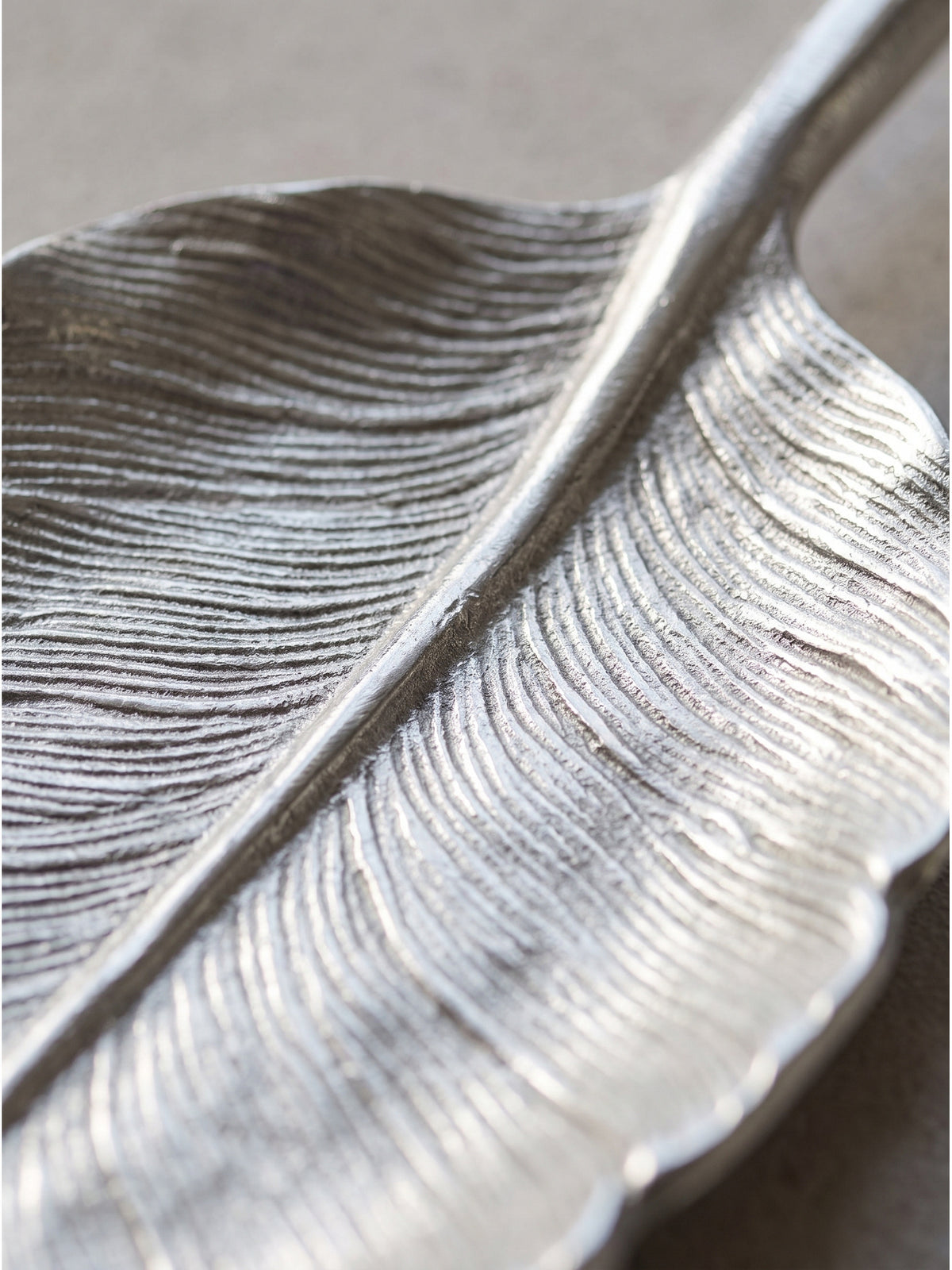 Nahaufnahme der lackierten Aluminium-Oberfläche, bei der die feinen Blattadern der Tischdeko-Schale vor einer Sichtbetonwand metallisch glänzen._