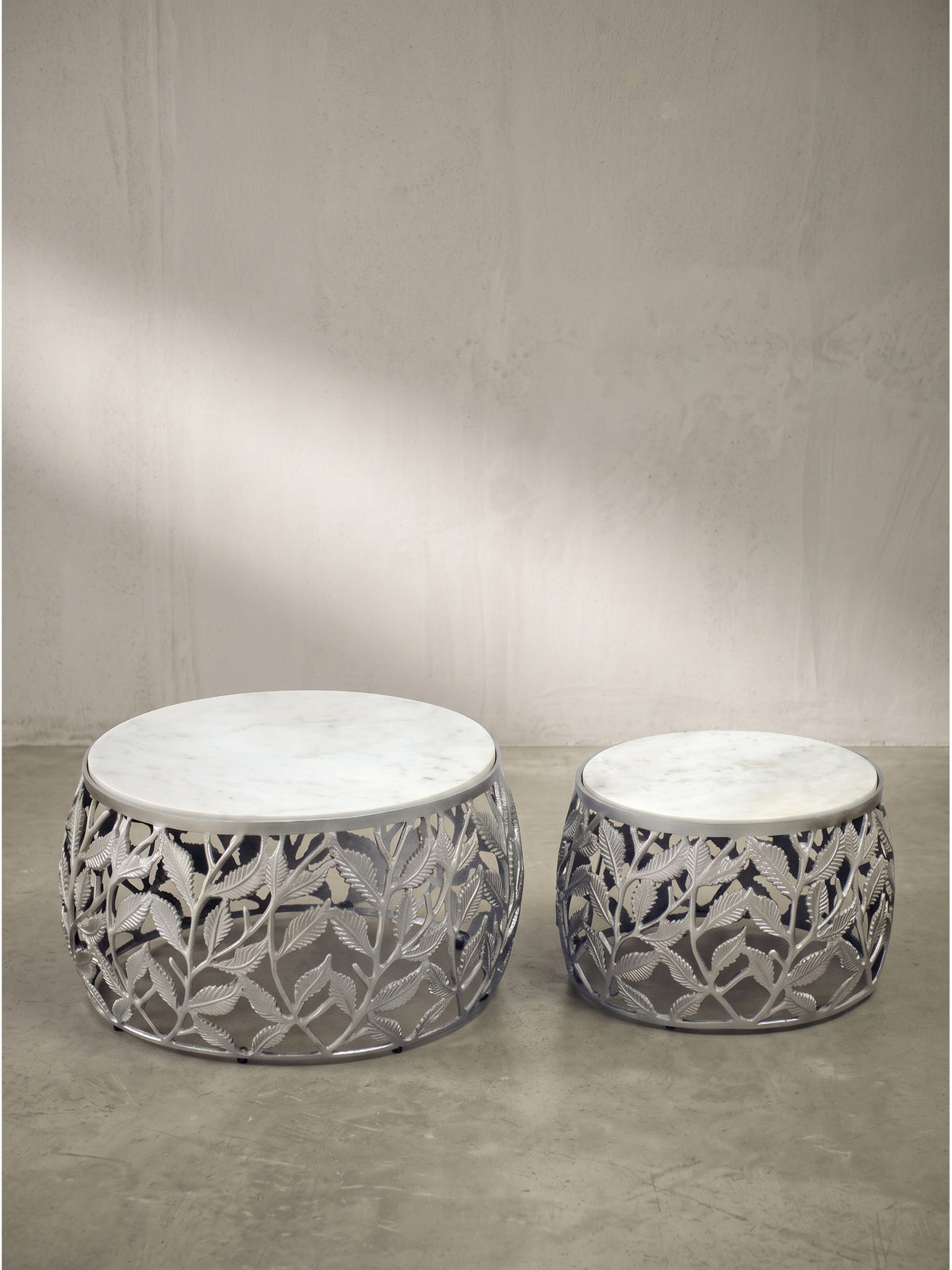 Frontalansicht des zweiteiligen Sofatisch-Sets aus kühlem Aluminium mit floralem Silbergestell und robusten, weißen Marmorplatten vor einem neutralen, hellen Hintergrund.