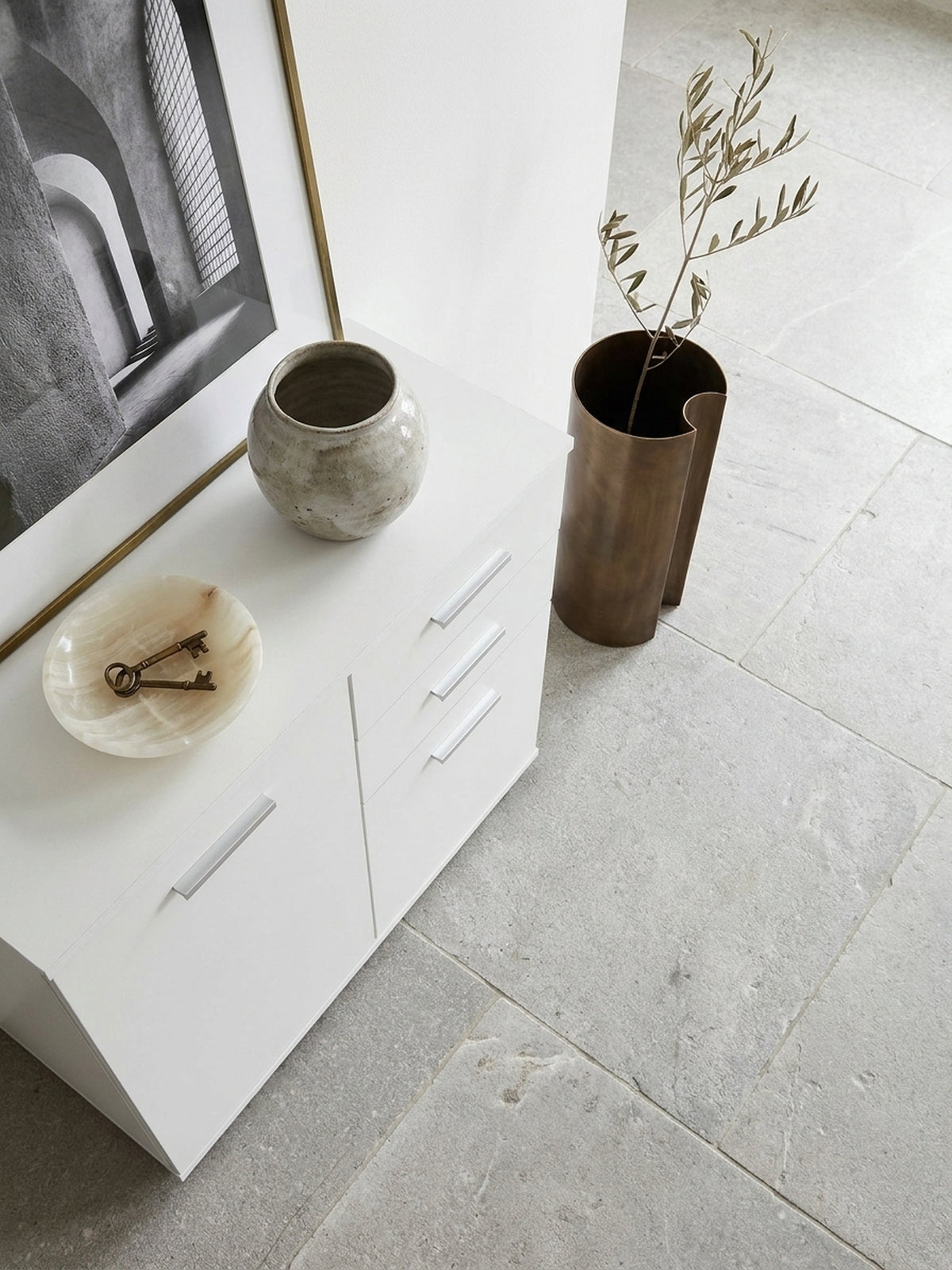 Kompakter weißer Mehrzweckschrank mit asymmetrischer Frontaufteilung von oben fotografiert auf großformatigen hellgrauen Kalksteinfliesen._
