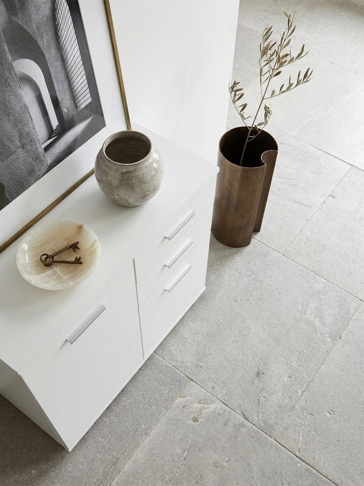 Kompakter weißer Mehrzweckschrank mit asymmetrischer Frontaufteilung von oben fotografiert auf großformatigen hellgrauen Kalksteinfliesen._