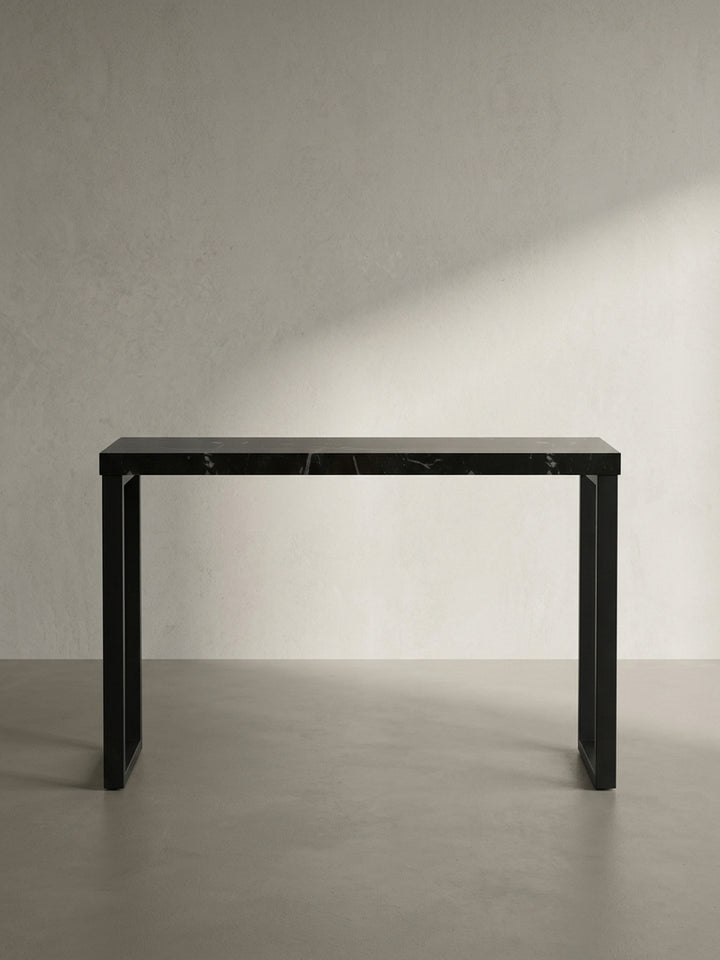 Schräge 3/4-Ansicht des Design-Ablagetisches, die die 5 cm dicke Tischplatte in Hochglanz-Schwarz und die geometrische Form des Gestells zeigt._