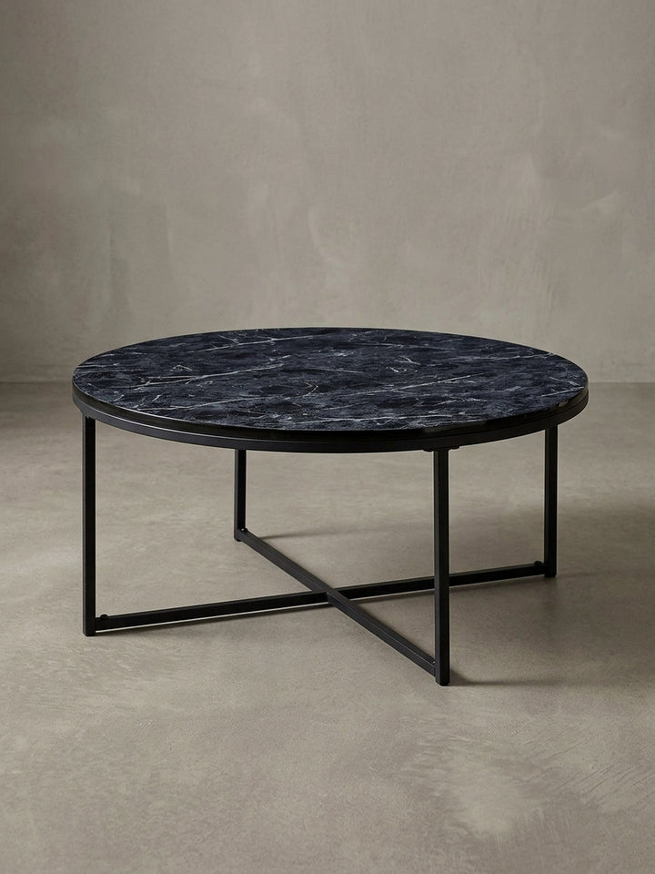 Design-Couchtisch in 3/4-Ansicht, die runde schwarze Marmor-Optik Platte und das gekreuzte Stahlgestell betonend, ideal für das moderne Wohnzimmer._