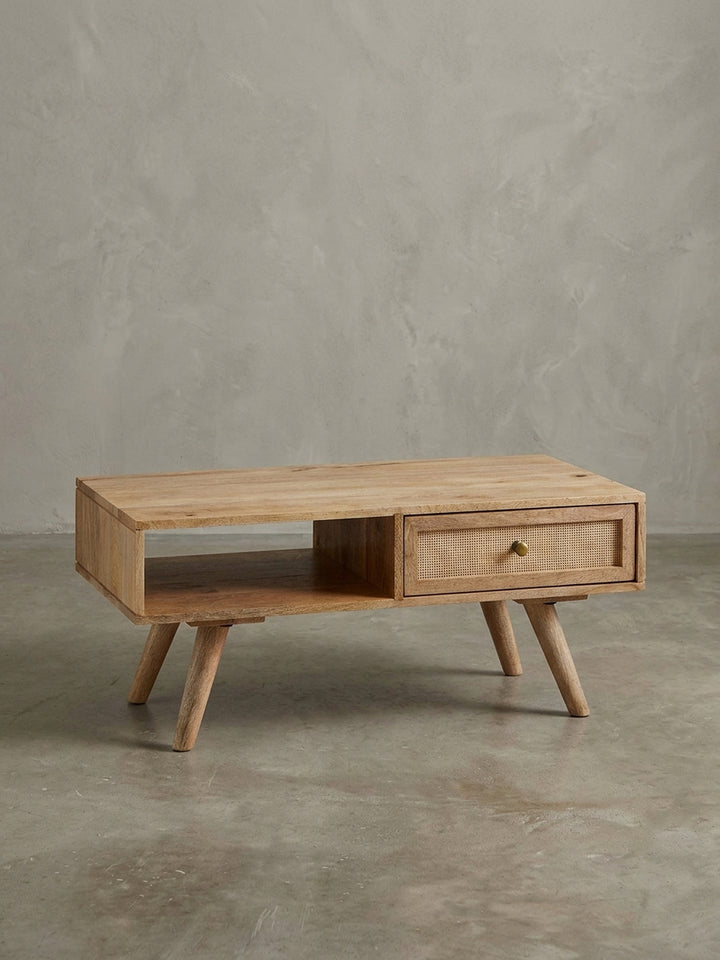 Design-Couchtisch in 3/4-Ansicht, der die asymmetrische Anordnung von Schublade mit Rattan und offenem Staufach aus massivem Mangoholz hervorhebt._