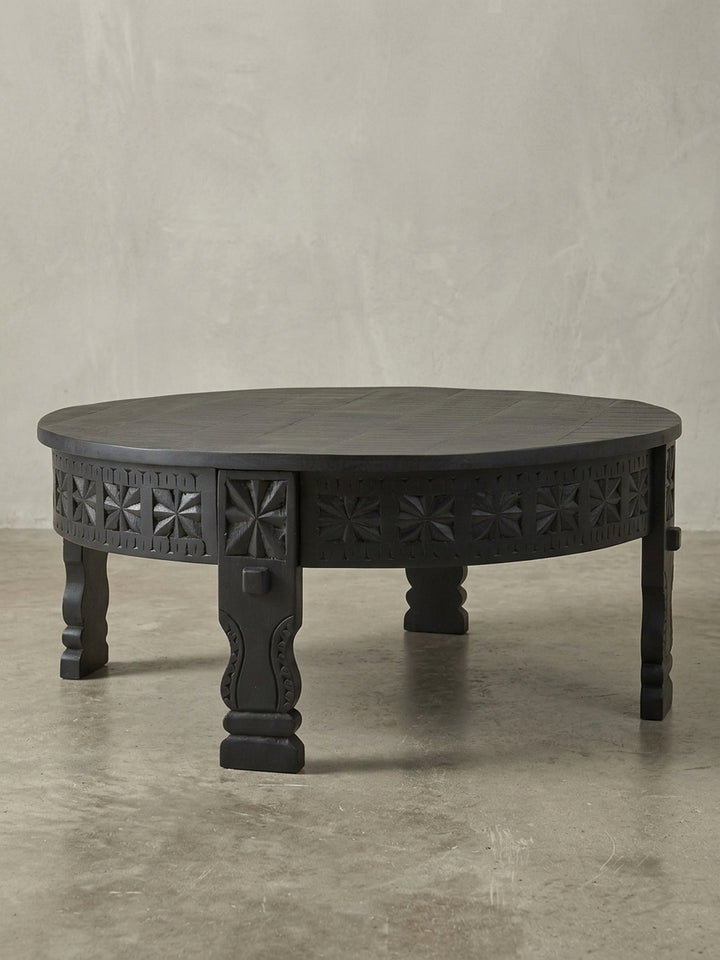 Dreiviertelansicht des massiven runden Sofatischs, der die kunstvollen orientalischen Schnitzereien an der Zarge und den robusten Beinen aus schwarzem Mangoholz detailreich zeigt._