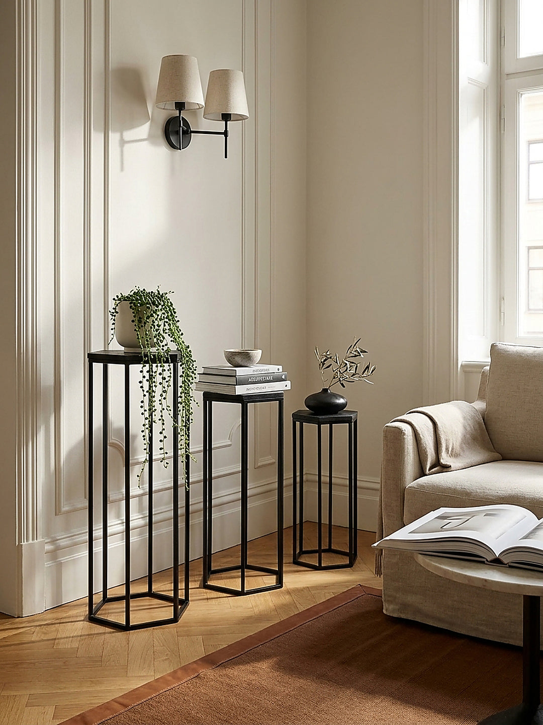 Minimalistisches Blumenständer Set in Schwarz kombiniert mit einem hellgrauen Leinensofa und einem tiefen architektonischen Wandpaneel._