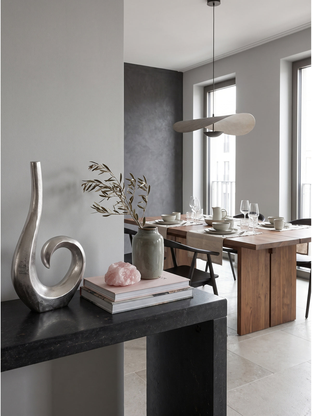 Standvase aus massivem Aluminium in Silber arrangiert mit einem hellgrauen Loungesessel auf kühlem Kalksteinboden._