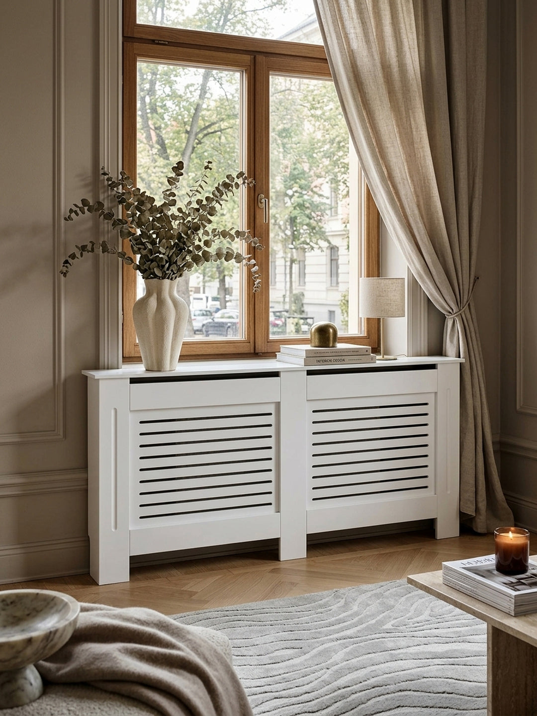 Elegante weiße Heizkörperverkleidung aus lackiertem MDF in einem klassisch-modernen Wohnzimmer. Matte Oberfläche, stilvoll als Konsole dekoriert unter einem großen Altbaufenster._