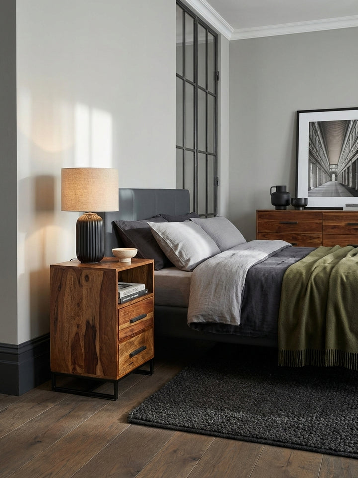 Eleganter Nachttisch aus massivem Sheeshamholz mit schwarzen Metallbeinen, perfekt passend zu einem hohen Boxspringbett in einem luxuriös eingerichteten, hellen Schlafzimmer._