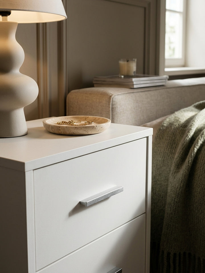 Nahaufnahme der weißen Melaminharzbeschichtung der Nachtkonsole. Die glatte pflegeleichte Oberfläche und die silbernen Griffe des Nachttischs zeigen puristisches Design._