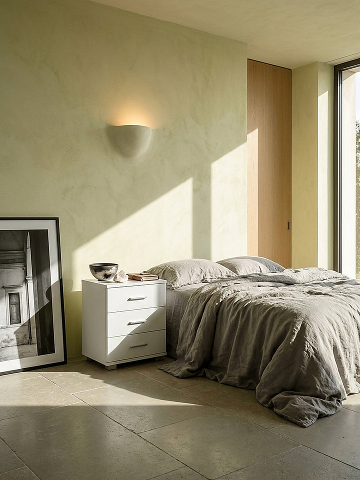 Luxuriöse weiße Nachtkonsole mit drei Schubladen neben einem grauen Leinenbett im minimalistischen Schlafzimmer mit edlem Kalksteinboden._