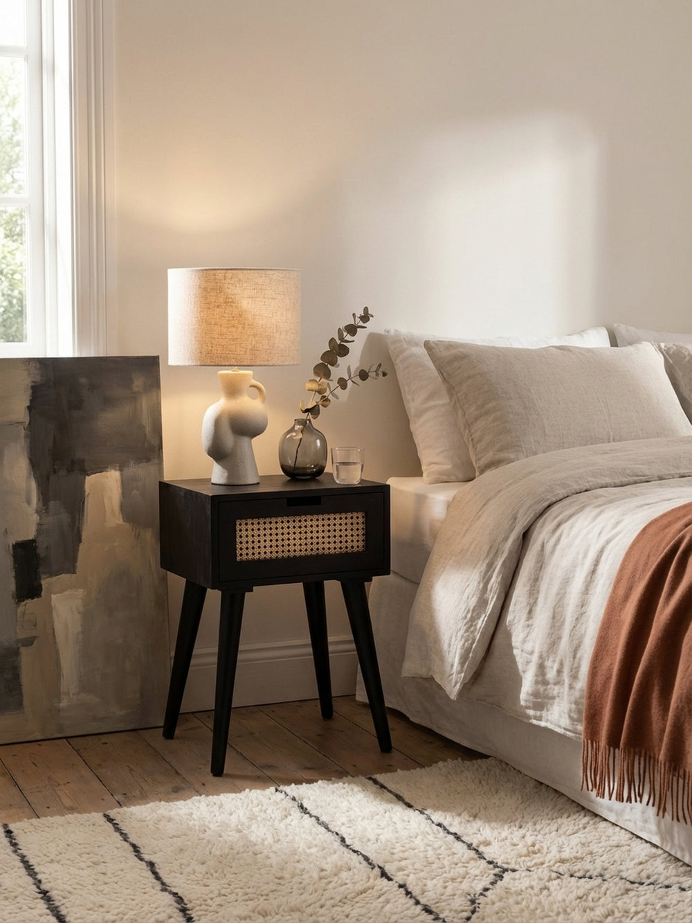 Schwarze Nachtkommode im Mid-Century-Stil kombiniert mit einem Bett mit hellem Stoffkopfteil und einer terrakottafarbenen Wolldecke für einen warmen Kontrast._