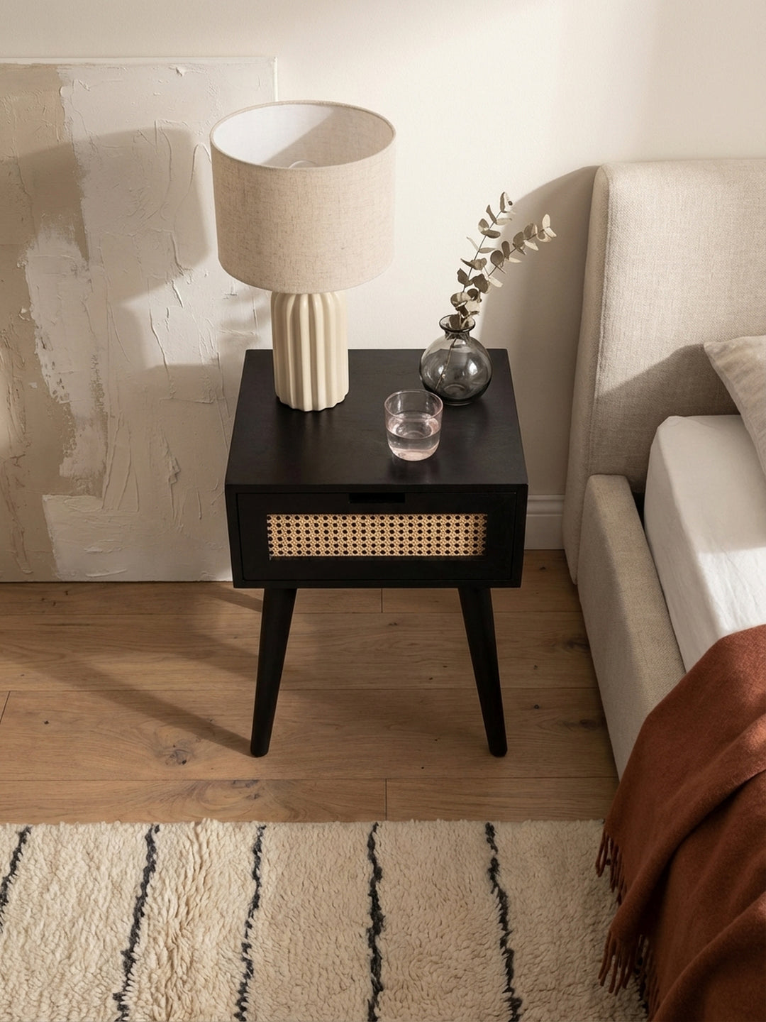 Draufsicht auf einen schwarzen Nachtschrank mit Rattangeflecht neben einem beigen Boxspringbett, platziert auf einem texturierten Beni Mrirt Teppich im Schlafzimmer._
