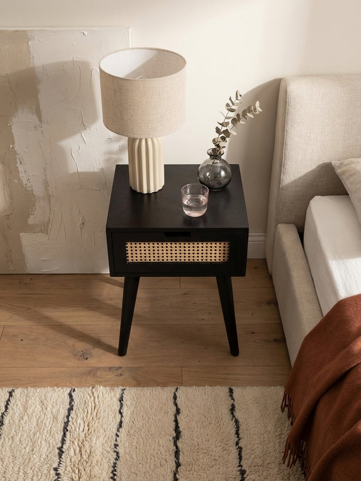 Draufsicht auf einen schwarzen Nachtschrank mit Rattangeflecht neben einem beigen Boxspringbett, platziert auf einem texturierten Beni Mrirt Teppich im Schlafzimmer._