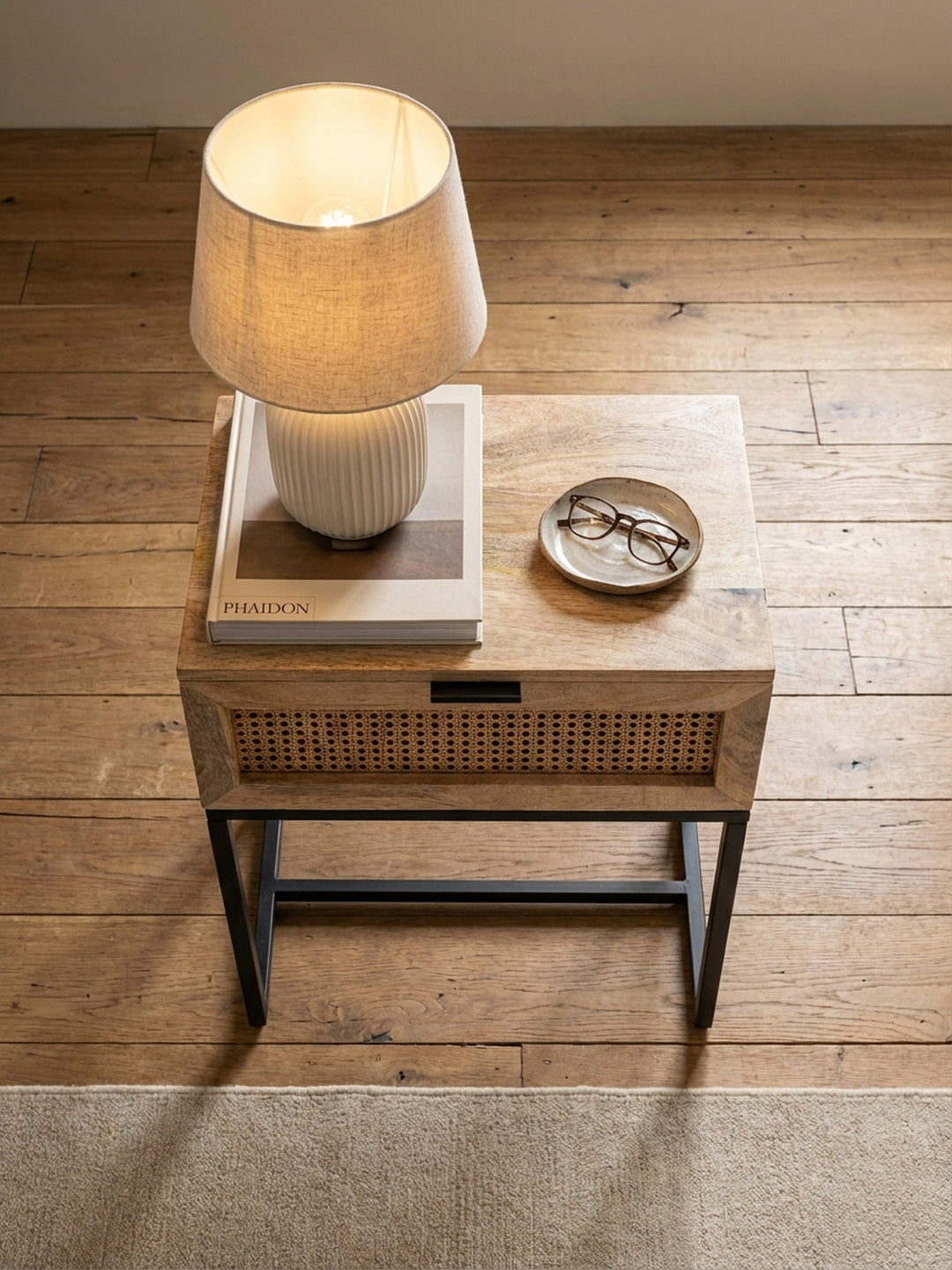 Draufsicht auf den Nachttisch aus Mangoholz neben einem Bett, platziert auf einem beigen Wollteppich mit Holzdielenboden._