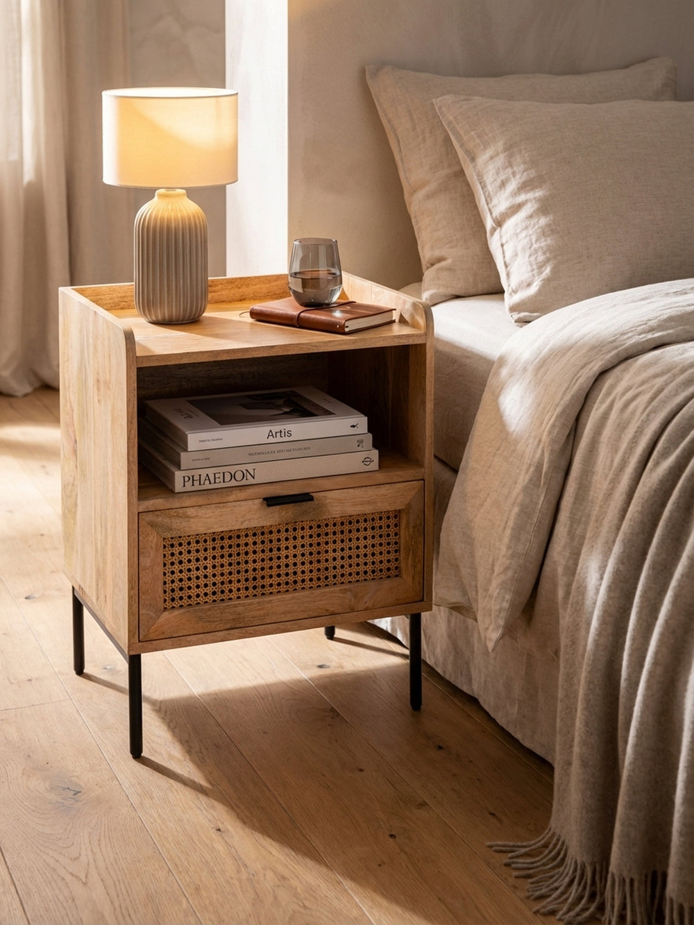 Die Nachtkommode mit Wiener Geflecht, dekoriert mit einer weißen Designervase, einem offenen Buch und einem Glas auf der Massivholzoberfläche._